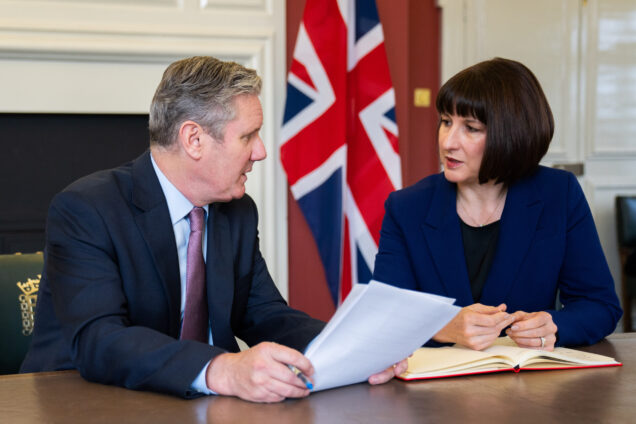 Keir Starmer and Rachel Reeves sat at a table, looking at each other in front of a Union flag