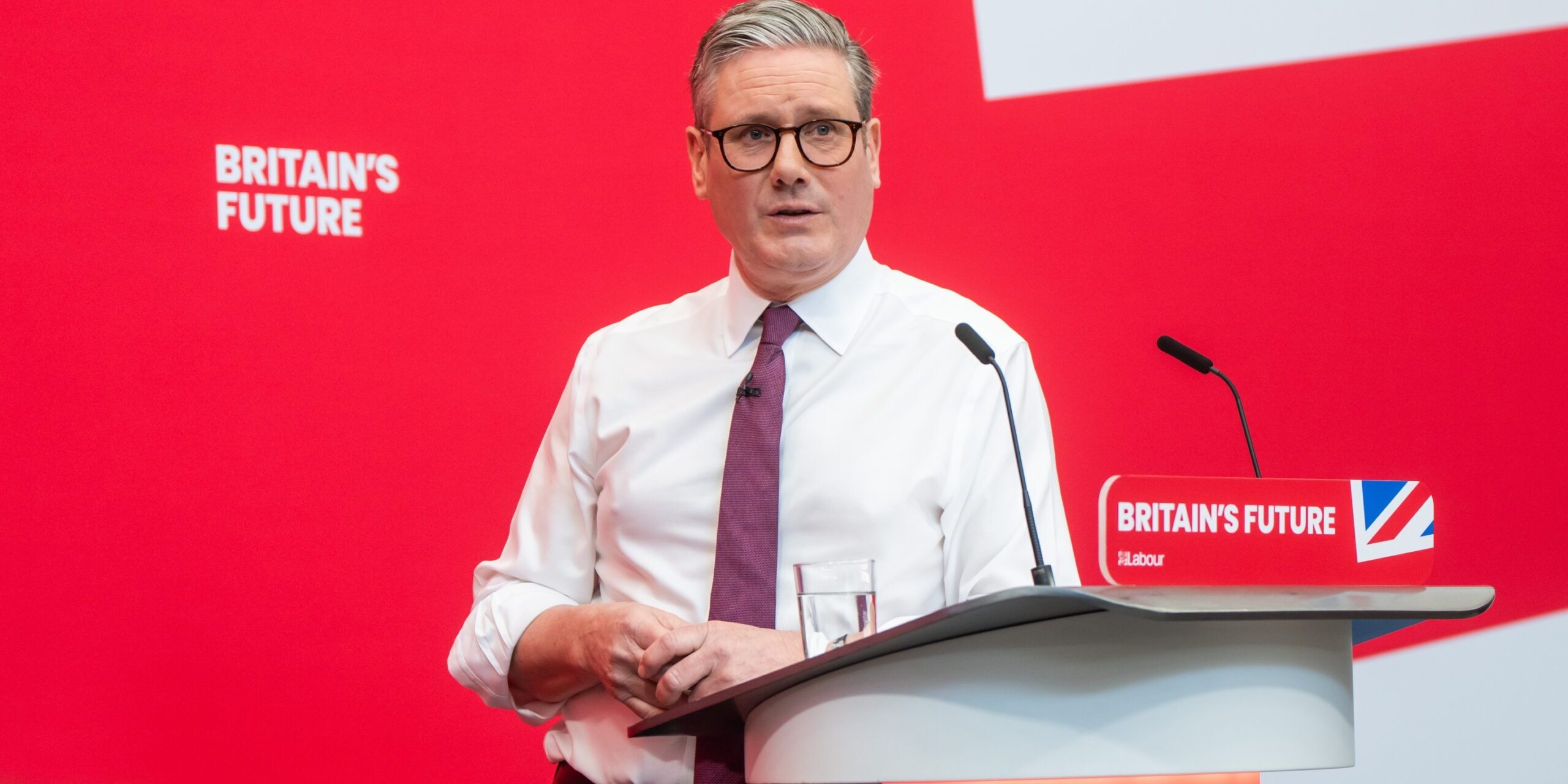 Keir Starmer standing at a podium in front of a red background with the phrase 'Britain's Future' written on it