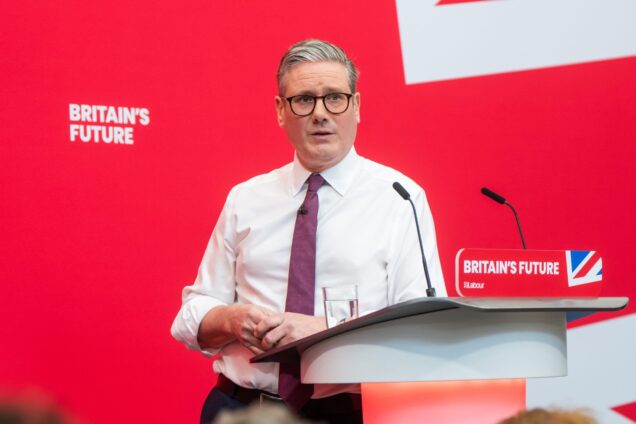 Keir Starmer standing at a podium in front of a red background with the phrase 'Britain's Future' written on it