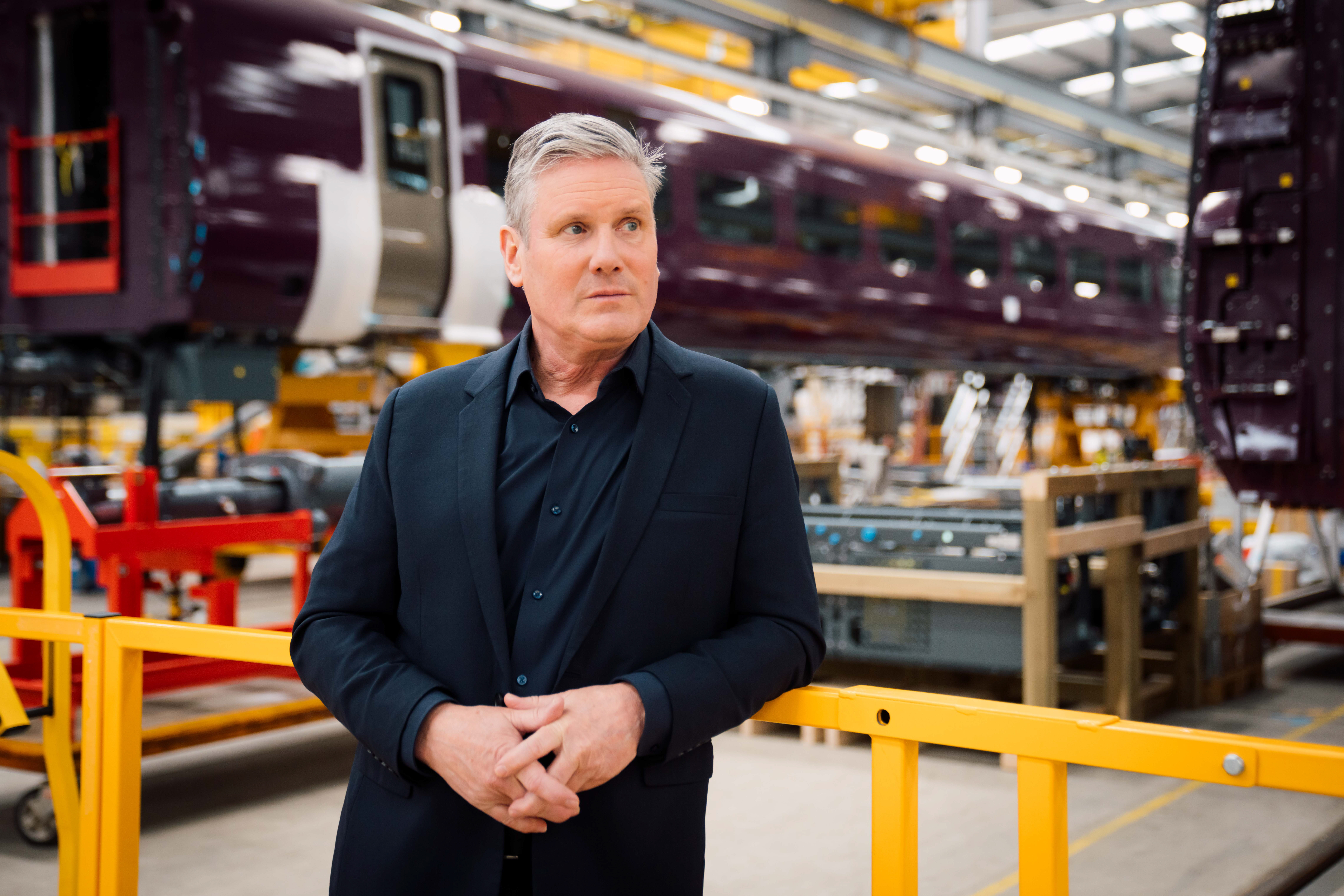 Keir Starmer stood in a factory in front of two train carriages, looking to the right of the camera shot