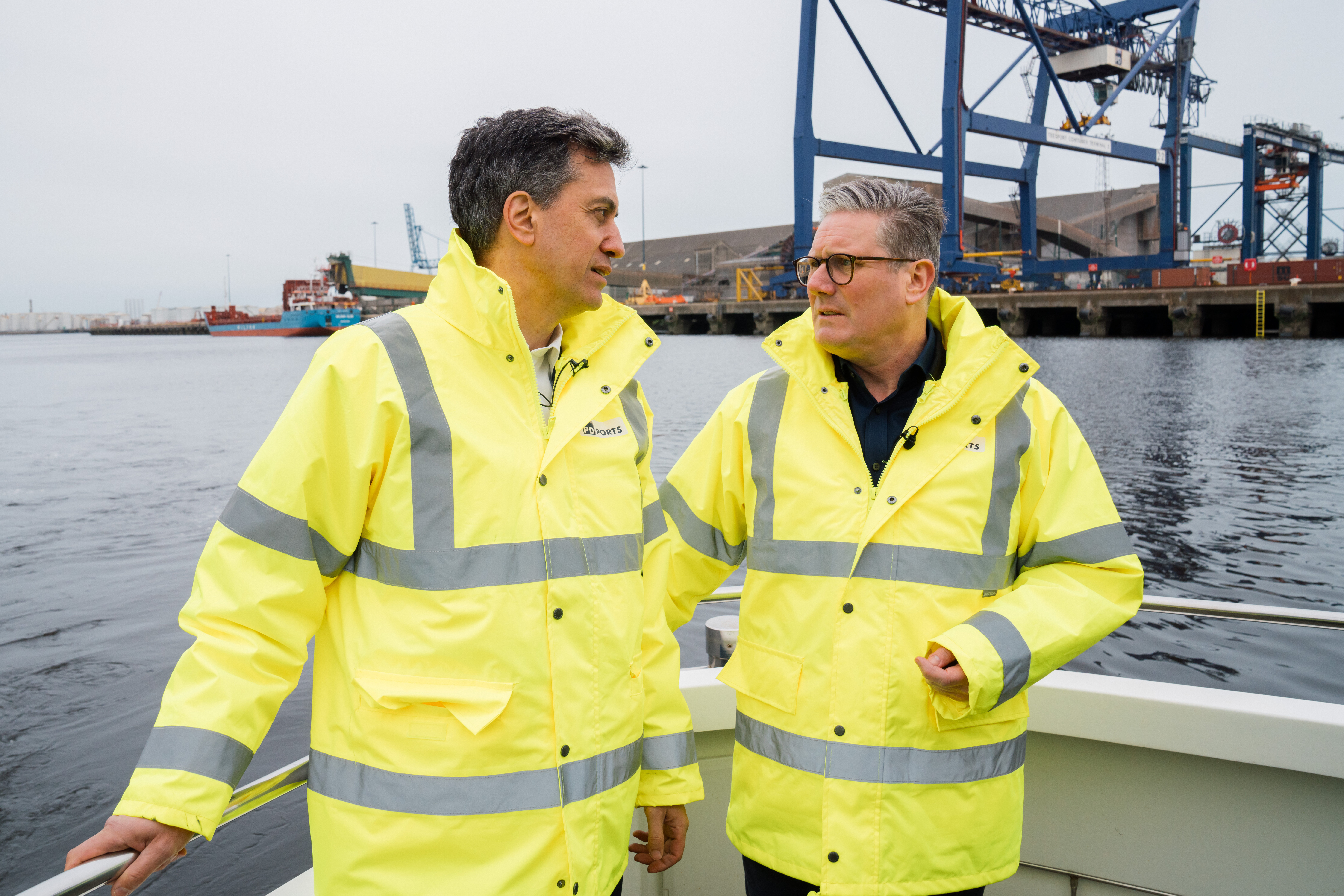 Keir Starmer and Ed Miliband talking to each other at a port, wearing high visibility yellow coats.