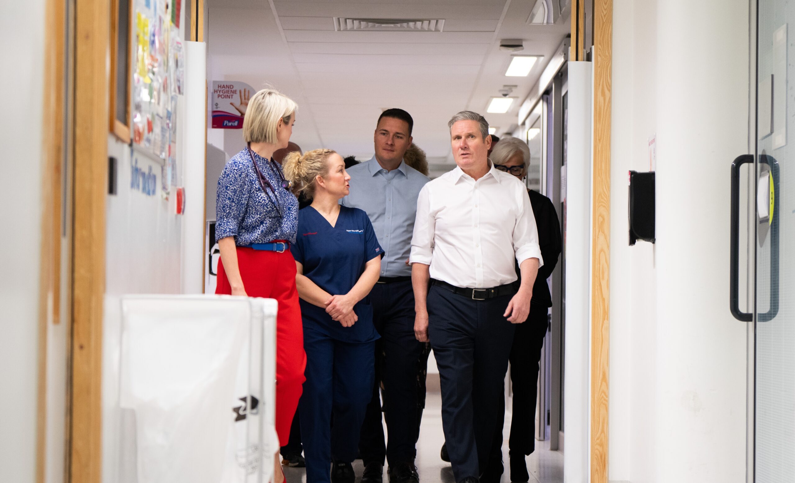 Keir Starmer walking down a corridor in a hospital with Wes Streeting and NHS staff.