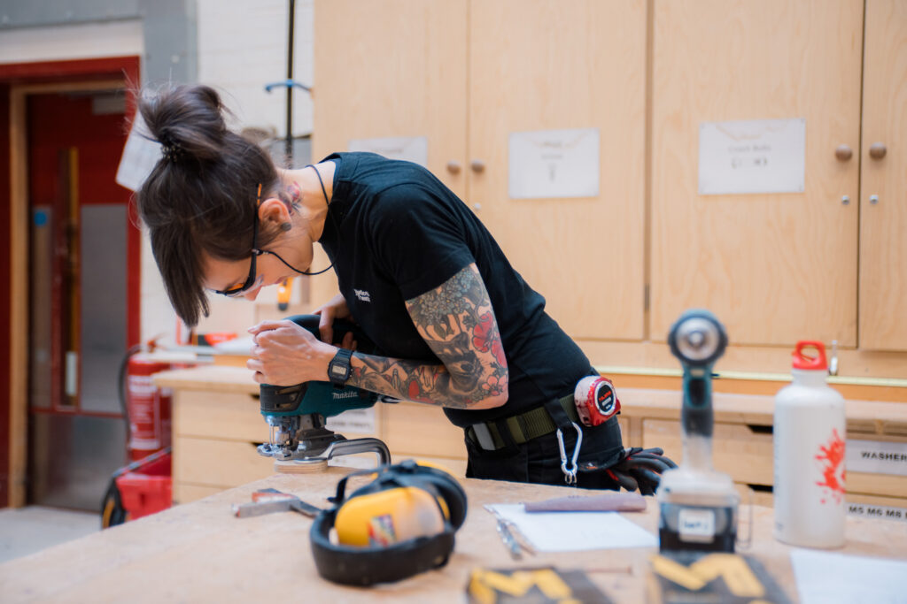 A woman does woodwork with an electric tool.