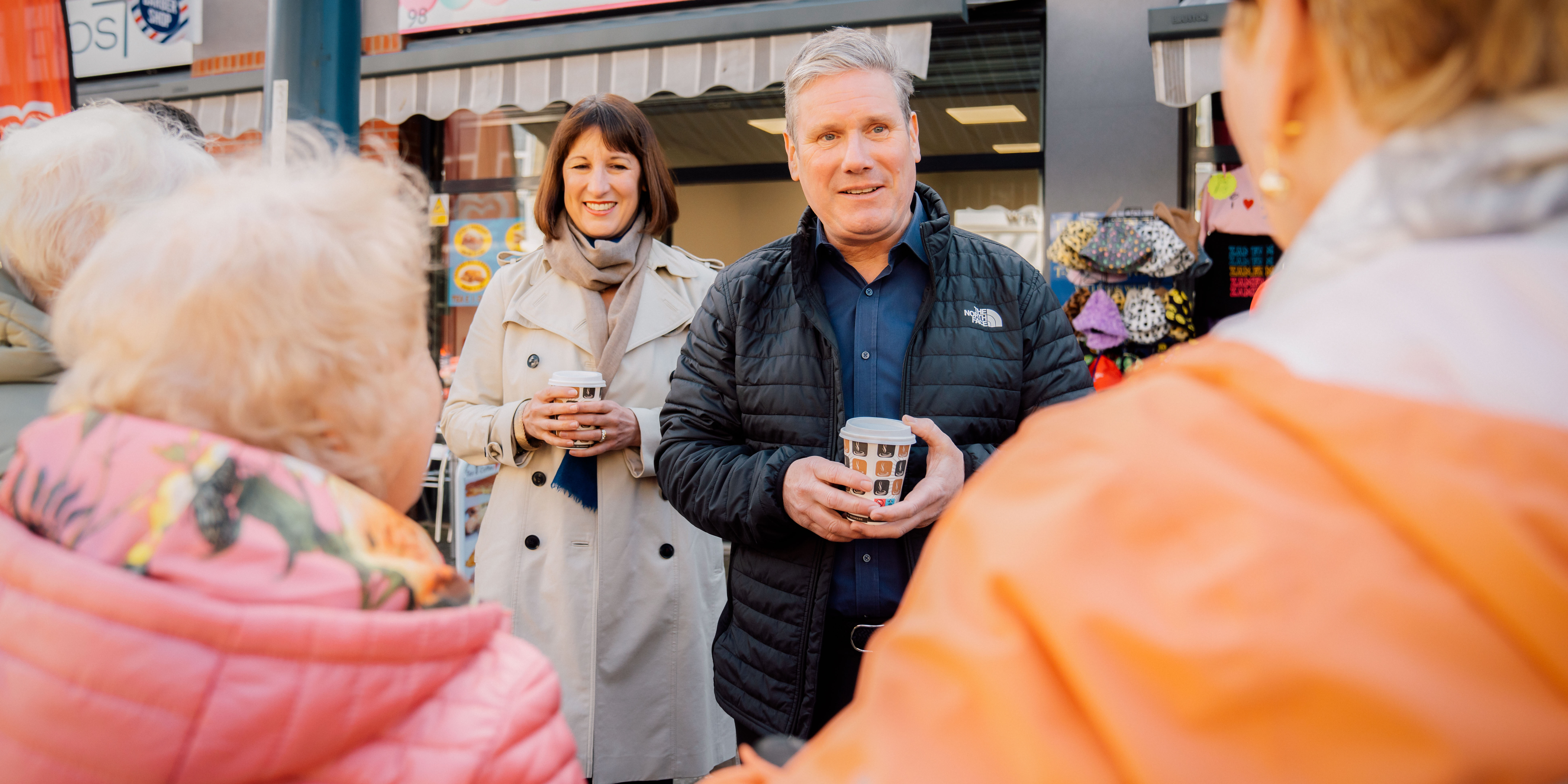 Photo of Keir Starmer and Rachel Reeves stood outside of a sweet shop, holding takeaway coffee cups and speaking to two women with their backs to the camera