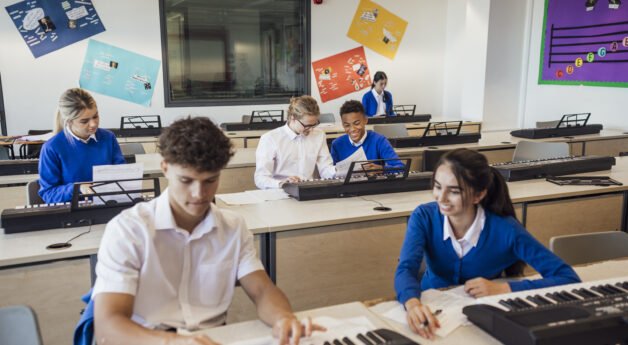 Teenage music students in a classroom learning how to play the keyboard together at a school. The students are wearing school uniforms.
