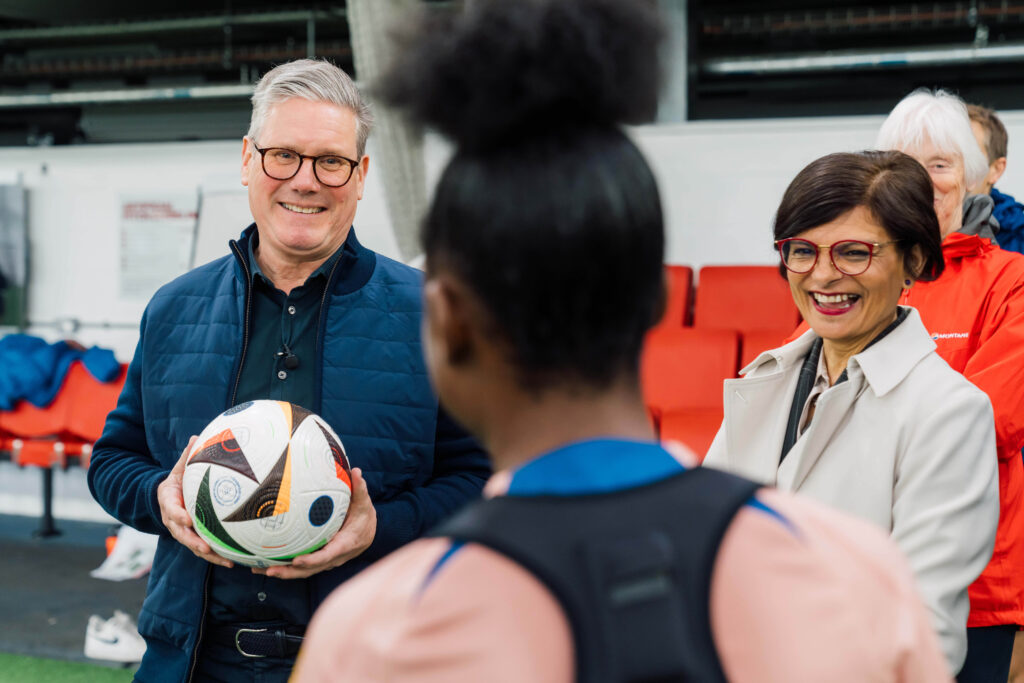 Keir Starmer and Thangam Debbonaire talk to a young woman on a football pitch. The Labour leader holds a ball.