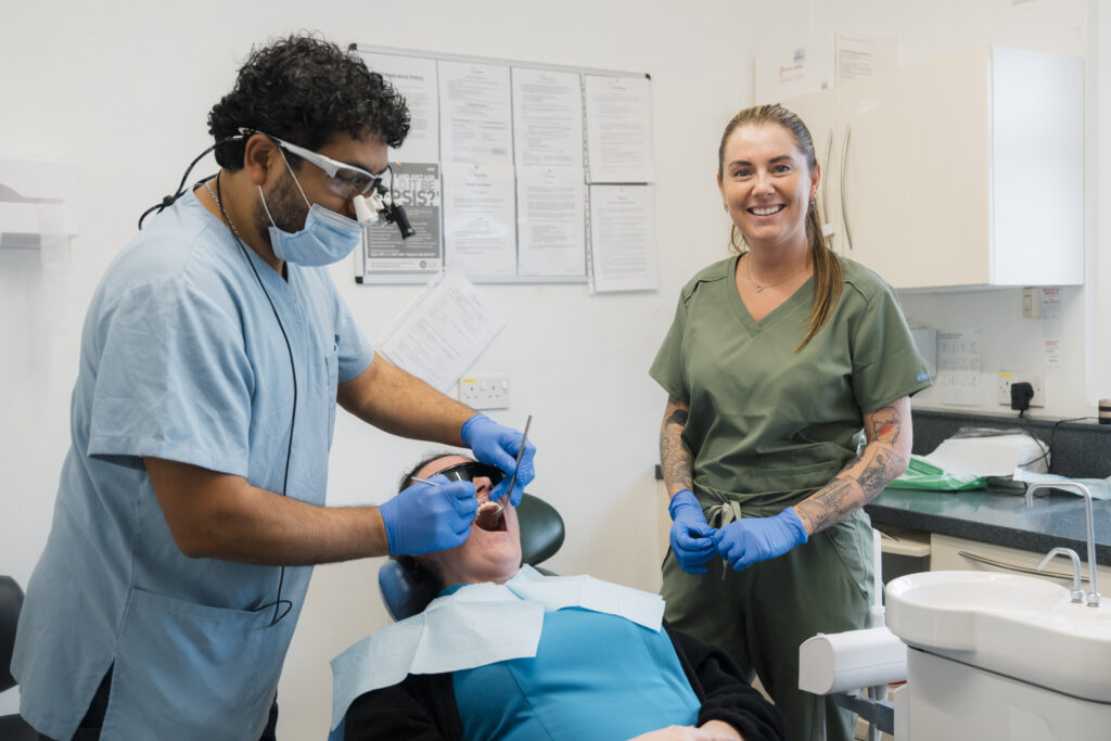 A dentist treats a seated patient with a support worker alongside.