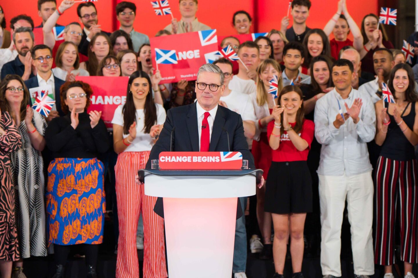 Keir Starmer at a lectern facing the camera, with supporters behind him holding placards reading 'Change', and small UK, Scottish and Welsh flags