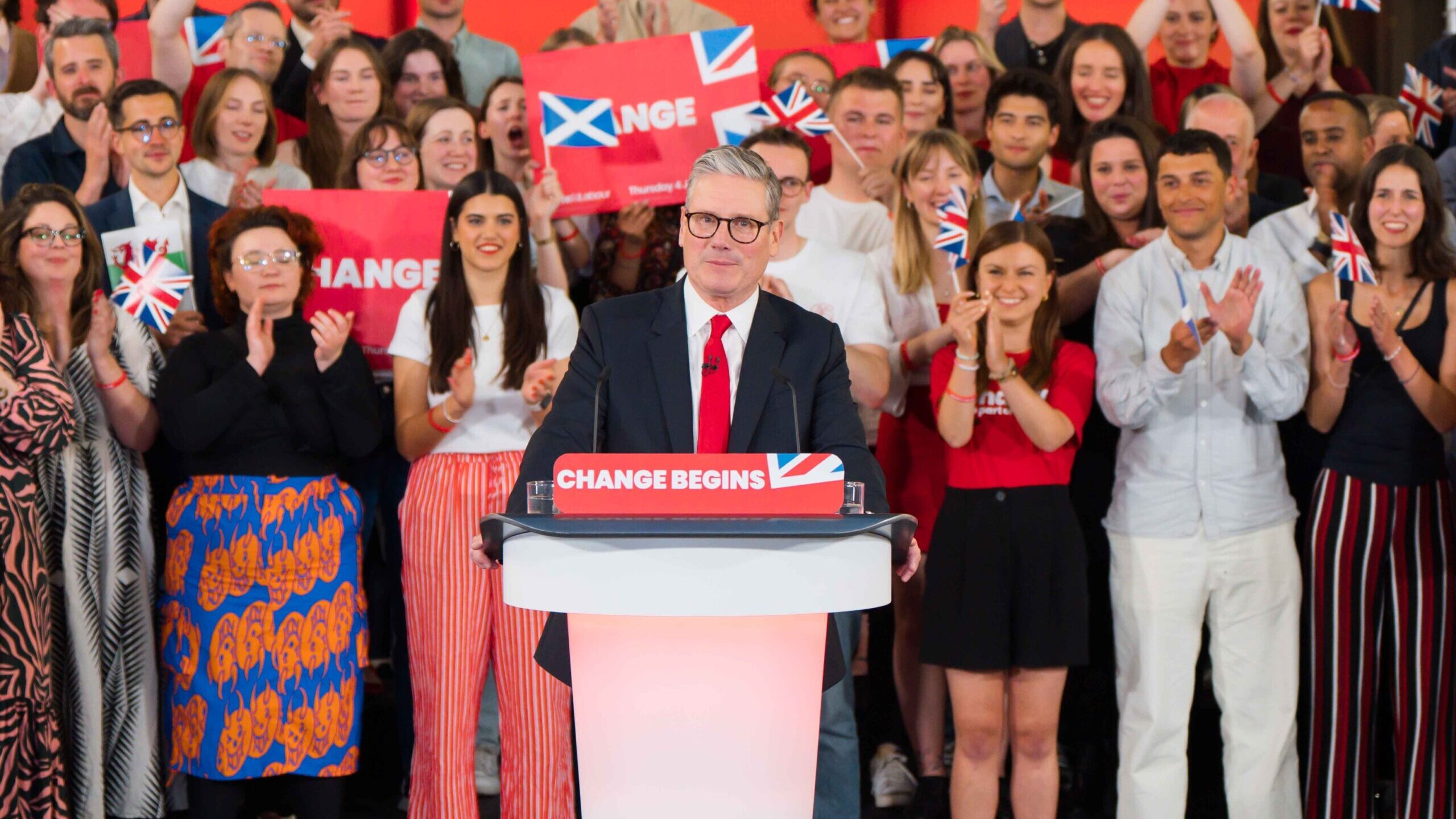 Keir Starmer at a lectern facing the camera, with supporters behind him holding placards reading 'Change', and small UK, Scottish and Welsh flags