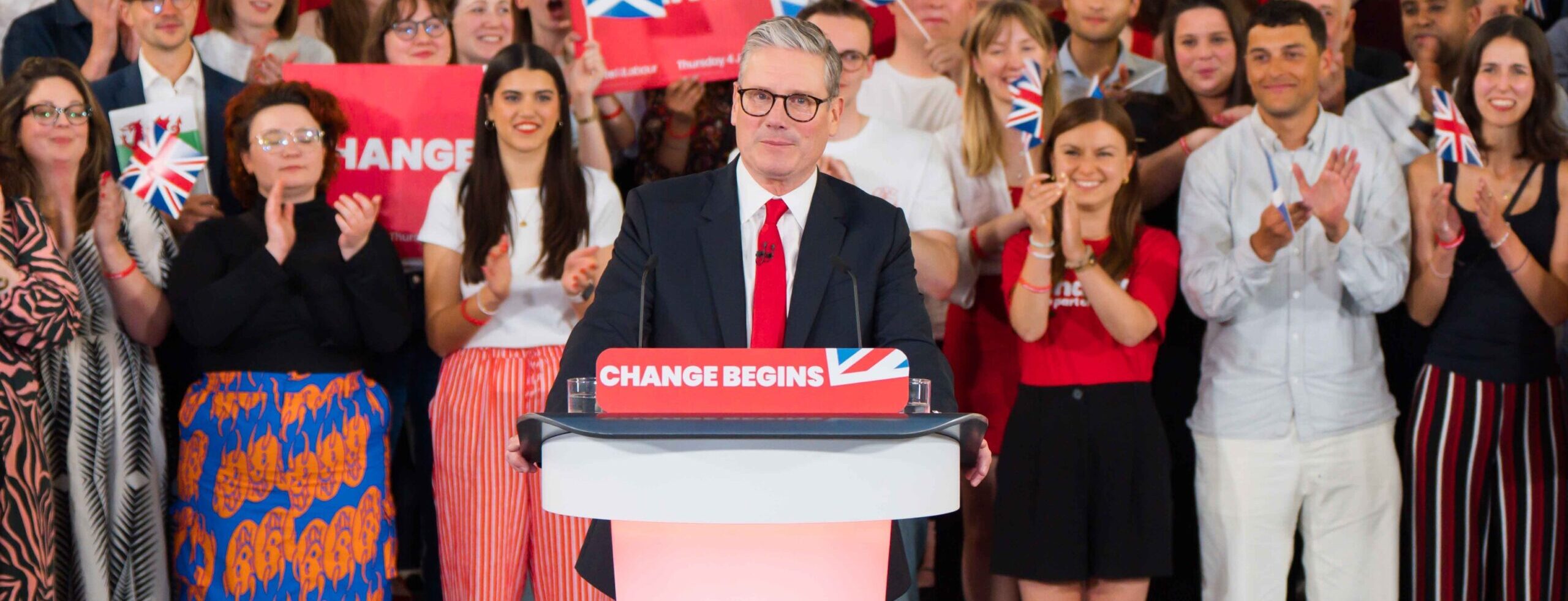 Keir Starmer at a lectern facing the camera, with supporters behind him holding placards reading 'Change', and small UK, Scottish and Welsh flags